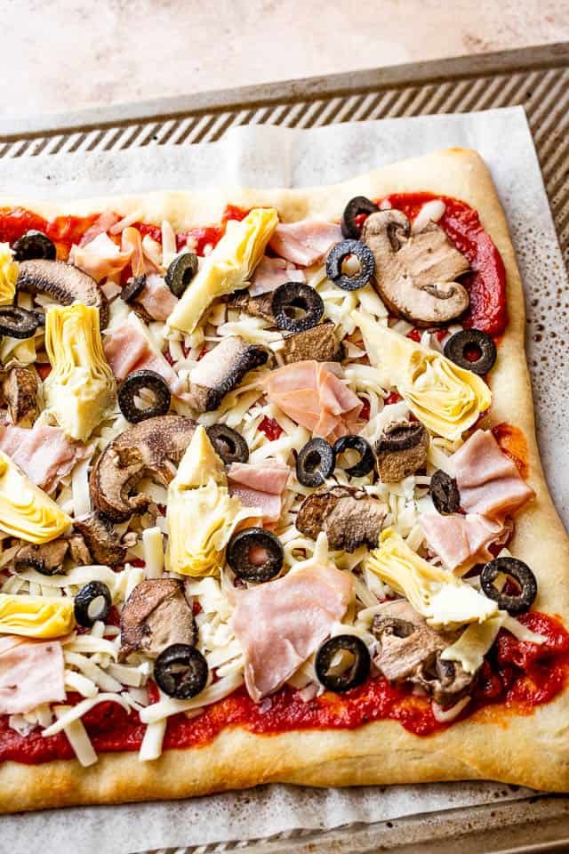 tomato sauce, cheese, artichoke hearts, olives, and ham on top of pizza crust prepared for grilling