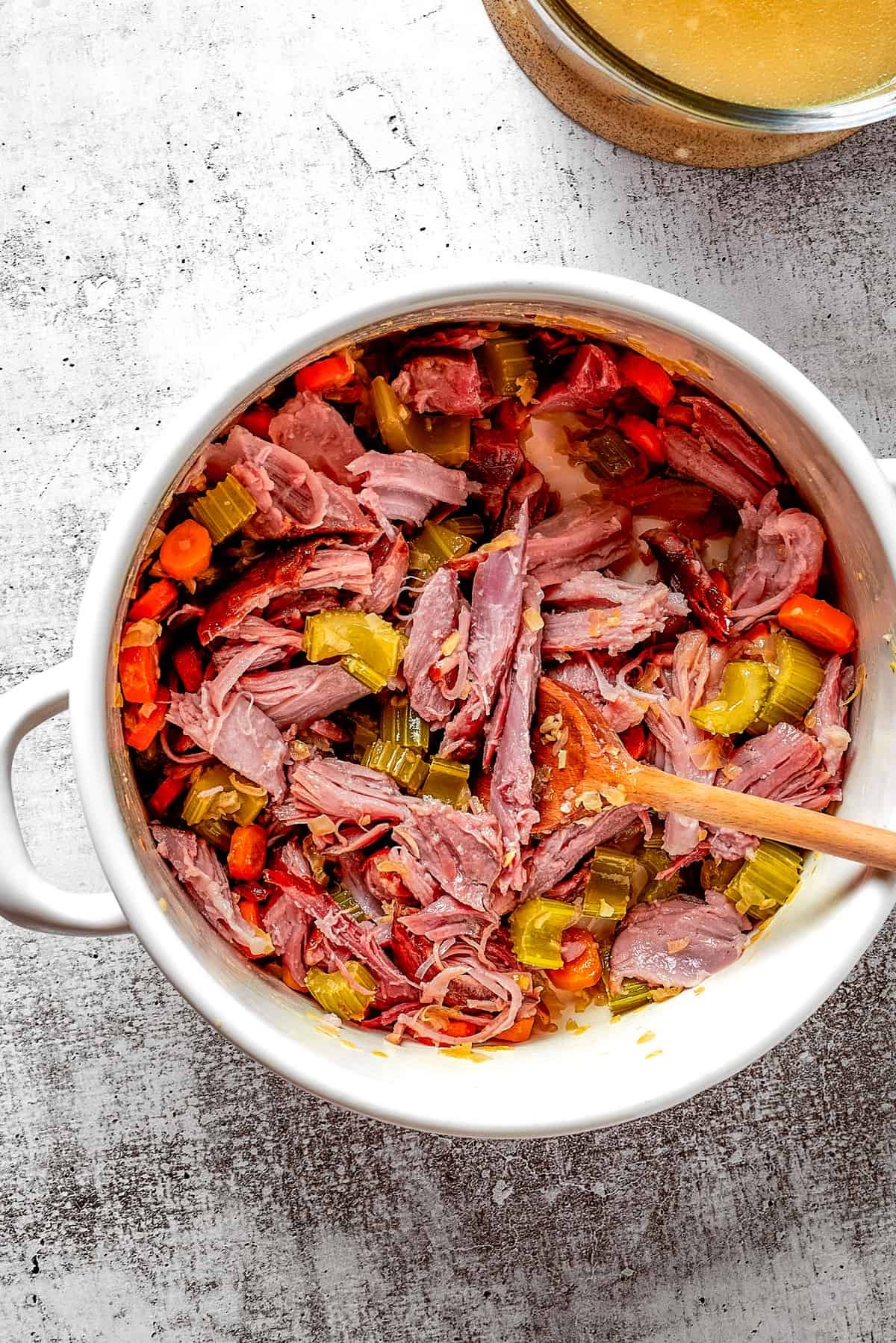 Overhead view of a dutch oven filled with veggies, ham, herbs, and a wooden spoon, next to a pyrex of broth.