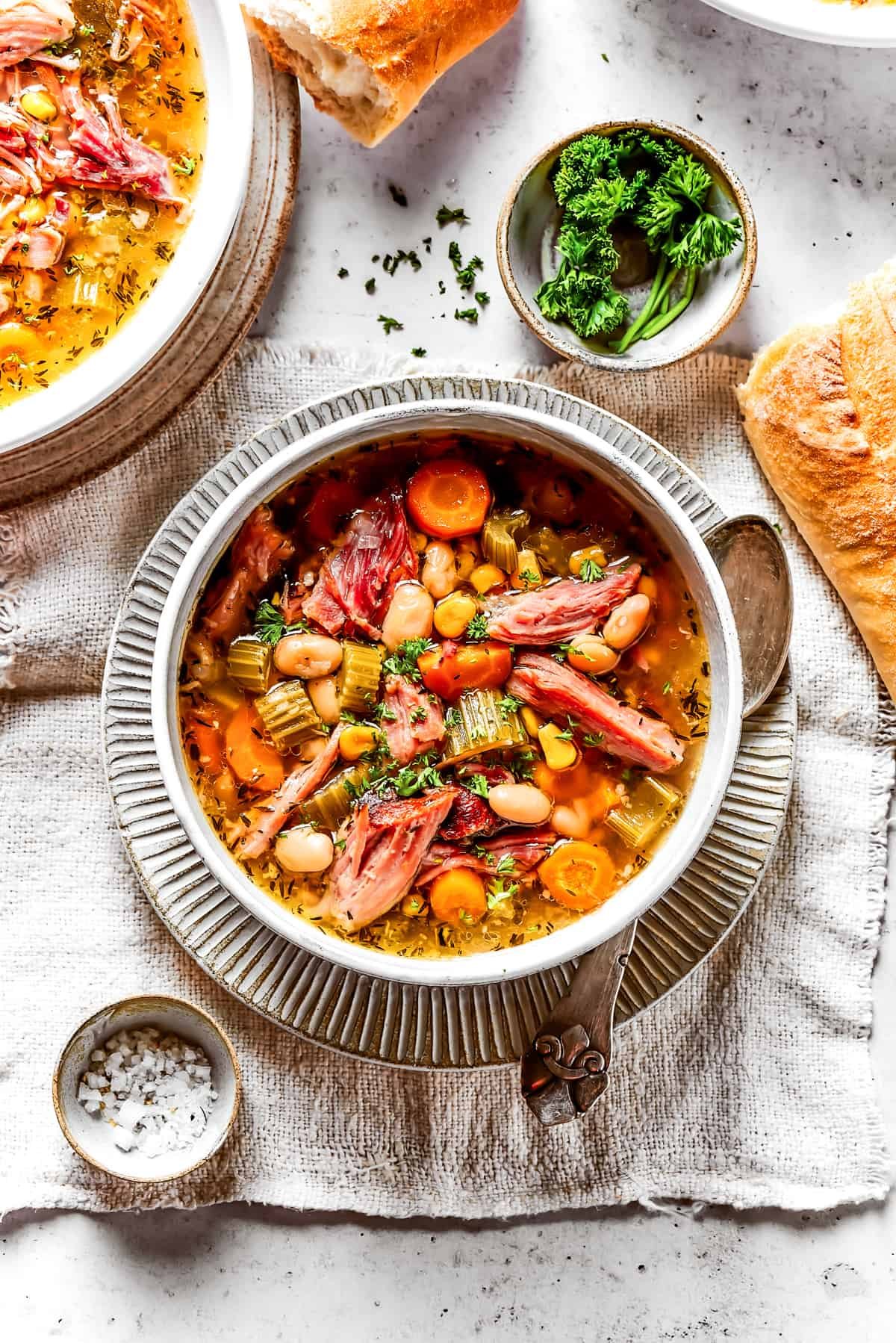 Overhead view of a bowl of ham bone soup with a spoon in it, next to a few pieces of baguette, another bowl of soup, and a bowl of salt.