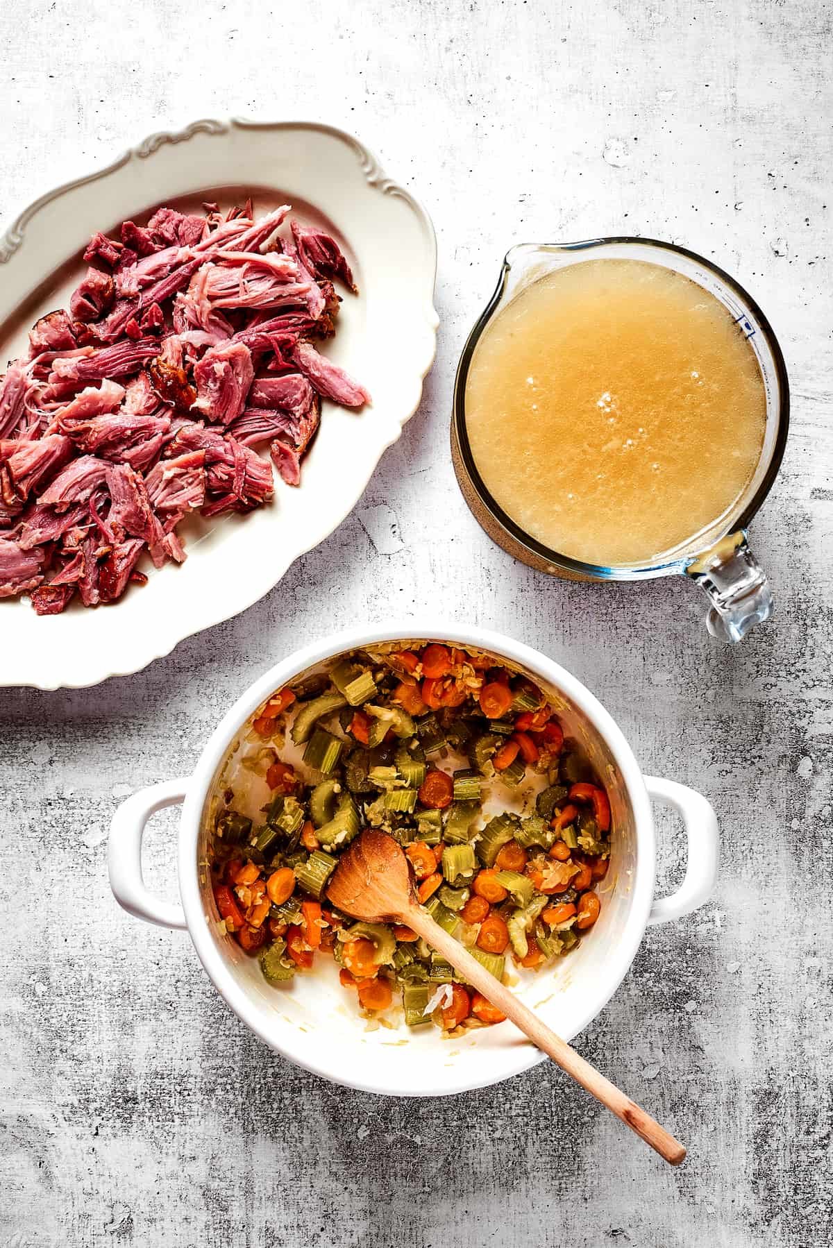 Overhead view of a dutch oven with onions, garlic, celery, carrots, and a wooden spoon, next to a pyrex of broth and a plate of shredded ham.
