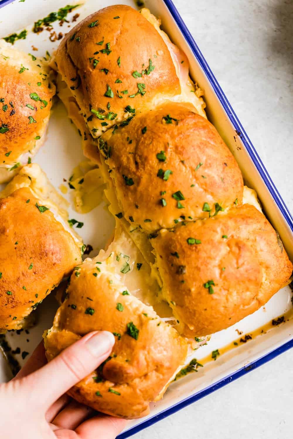 A Hand Grabbing a Ham and Cheese Slider From a Blue-Rimmed Pan