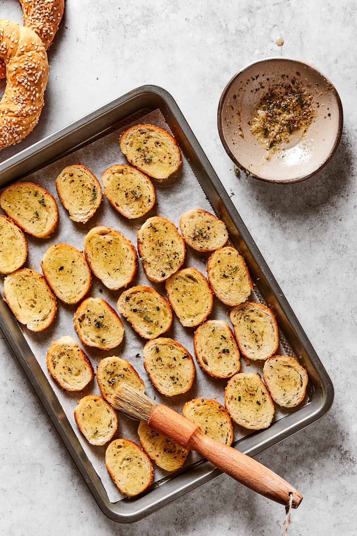 Bagel rounds lined up on a baking sheet, brushed with seasoned butter. A pastry brush is resting on the baking sheet, and dish of seasoned butter is next to it.