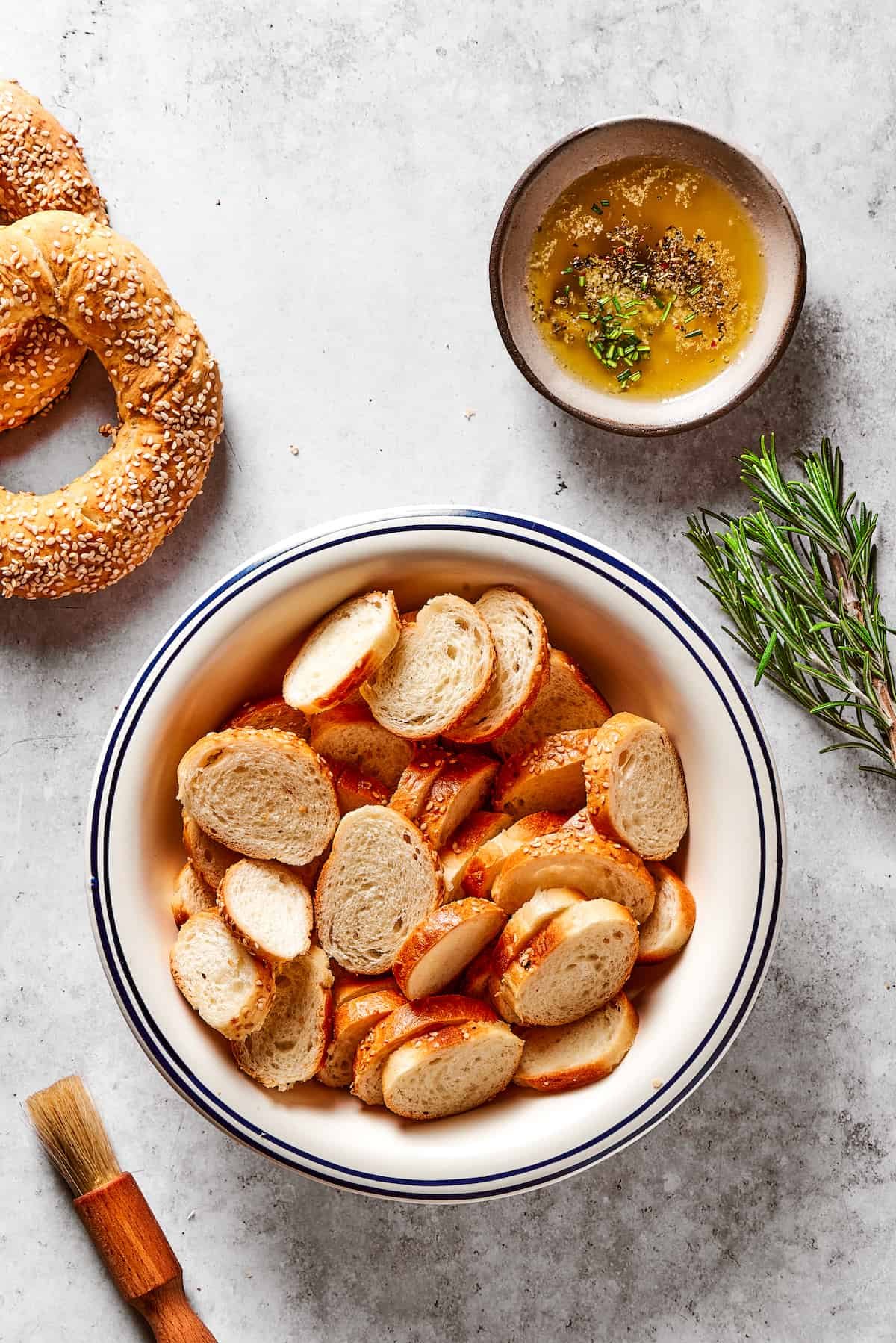 Sliced bagel rounds next to a dish of seasoned butter and a sprig of rosemary.