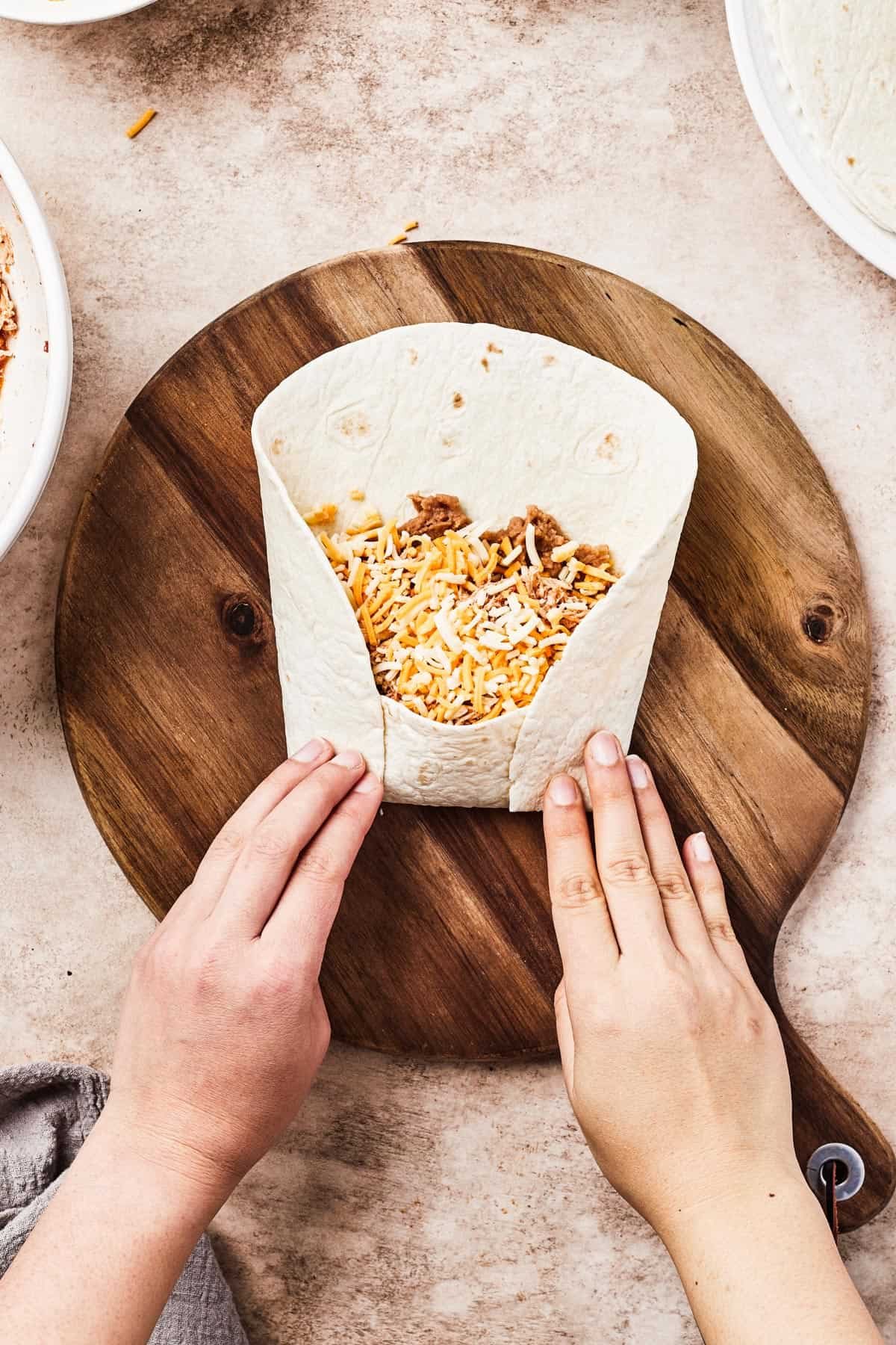 A woman folds the bottom and sides of a tortilla over the filling in the center of the tortilla.