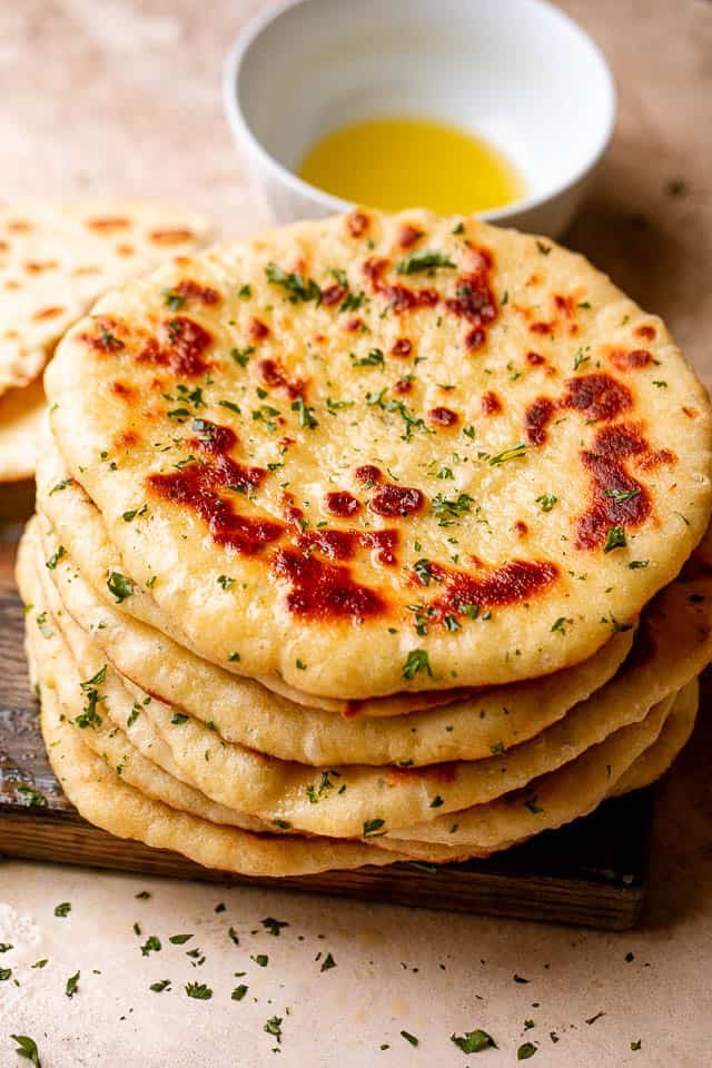 Top view of a stack of homemade flatbread on a wooden board next to a bowl of olive oil.