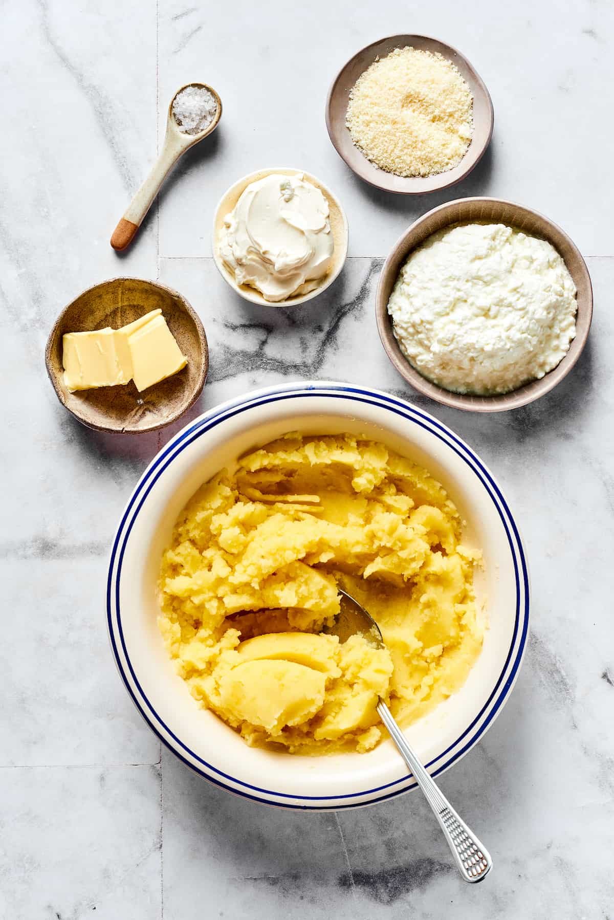 Mashed potatoes in a bowl with dishes of cottage cheese, sour cream, Parmesan, butter, and salt.