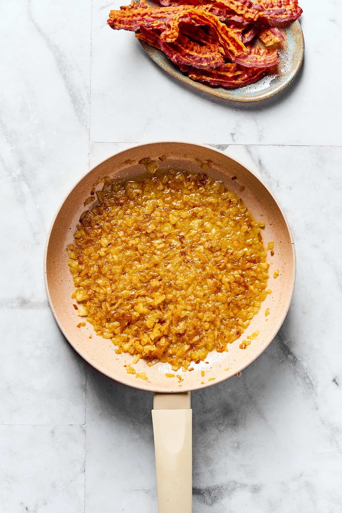 Caramelized onions and butter in a skillet. A plate of cooked bacon is nearby on the work surface.