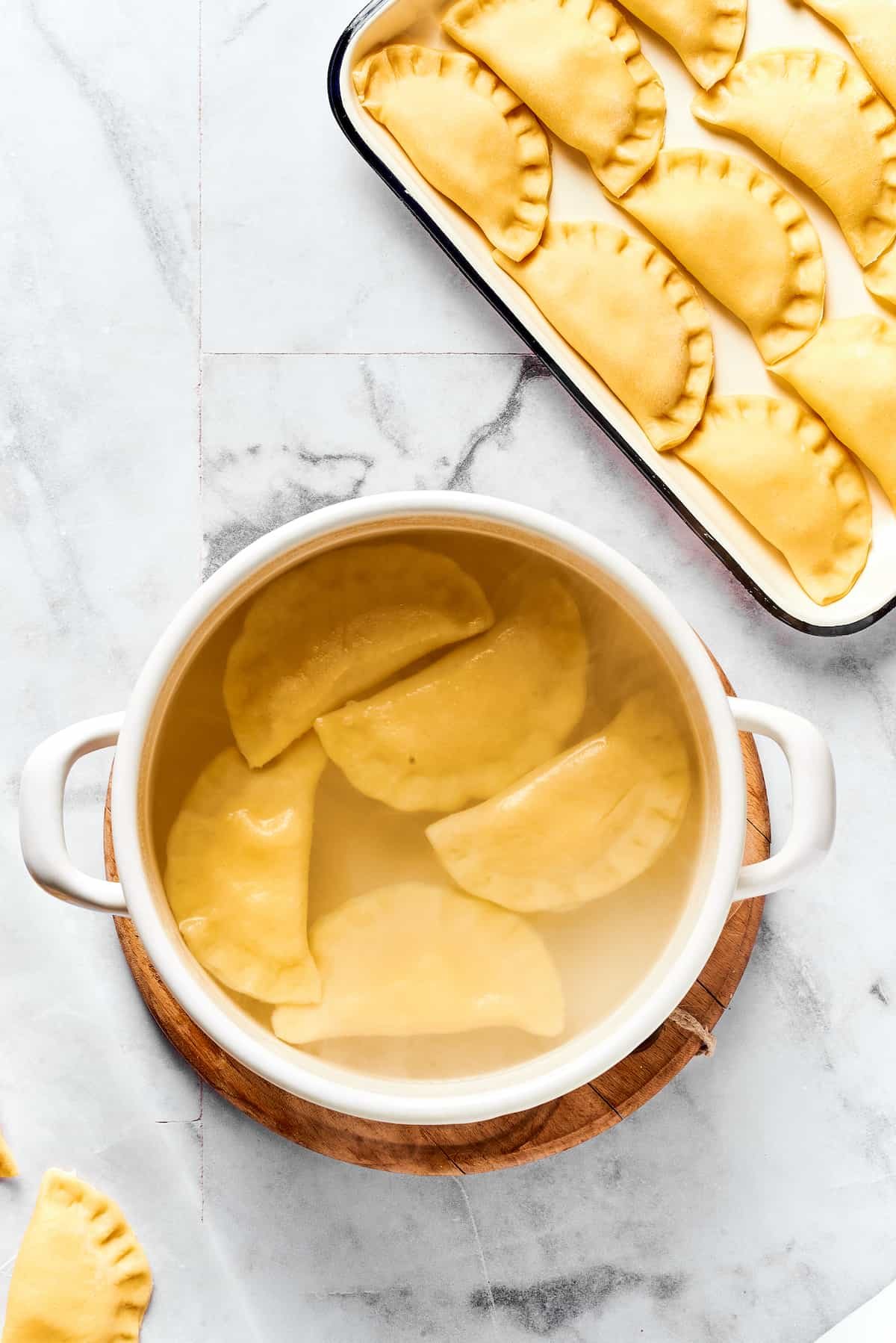 Pierogies boiling in a pot of water. A tray of uncooked pierogies is next to the pot.