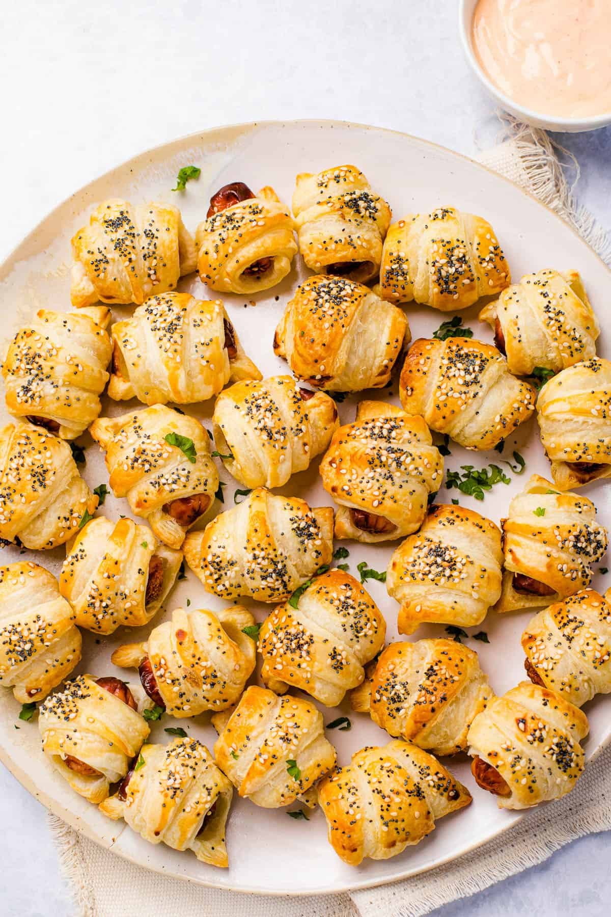 A serving platter holding about two dozen pigs in a blanket. A bowl of dipping sauce sits nearby.