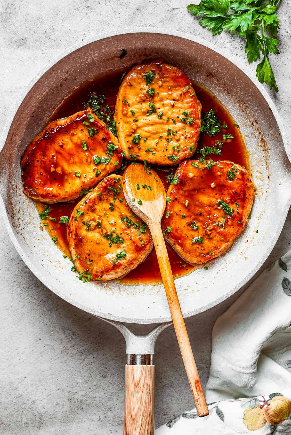 Four honey garlic pork chops cooking in a skillet with a wooden spoon stirring through.