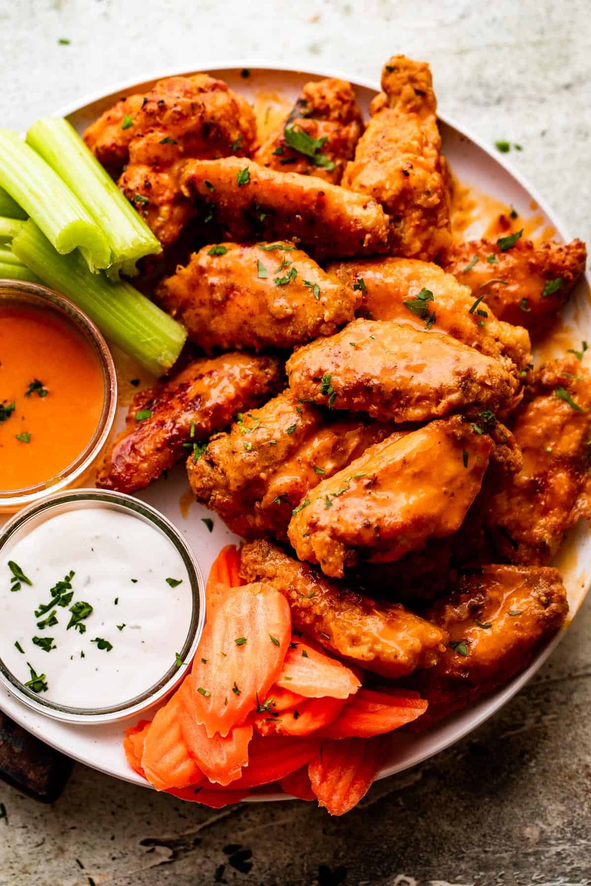 overhead shot of Mango Habanero Hot Wings arranged on a white serving plate with celery sticks and carrots set alongside the wings. Two Jars of hot sauce and ranch dipping sauce are also set on the plate.