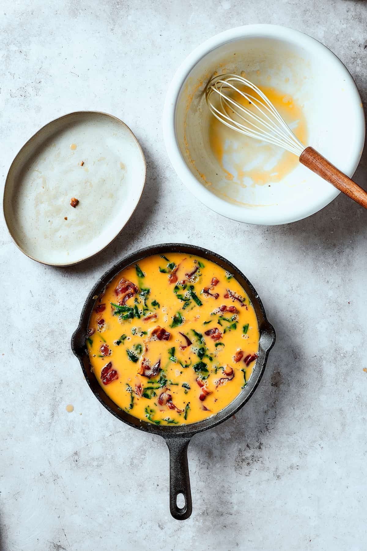 Pouring egg scramble over bacon, onion, spinach, and potatoes in a cast-iron pan.