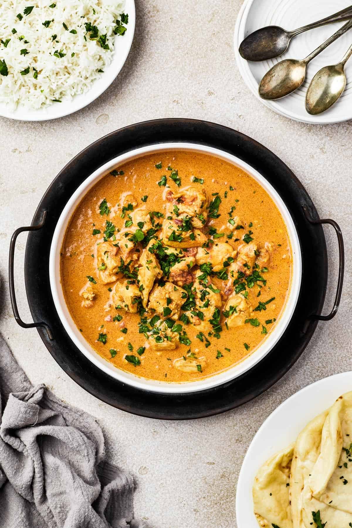 Overhead shot of a white enamel dish, resting on a cast-iron holder. The dish is filled with butter chicken, garnished with cilantro.