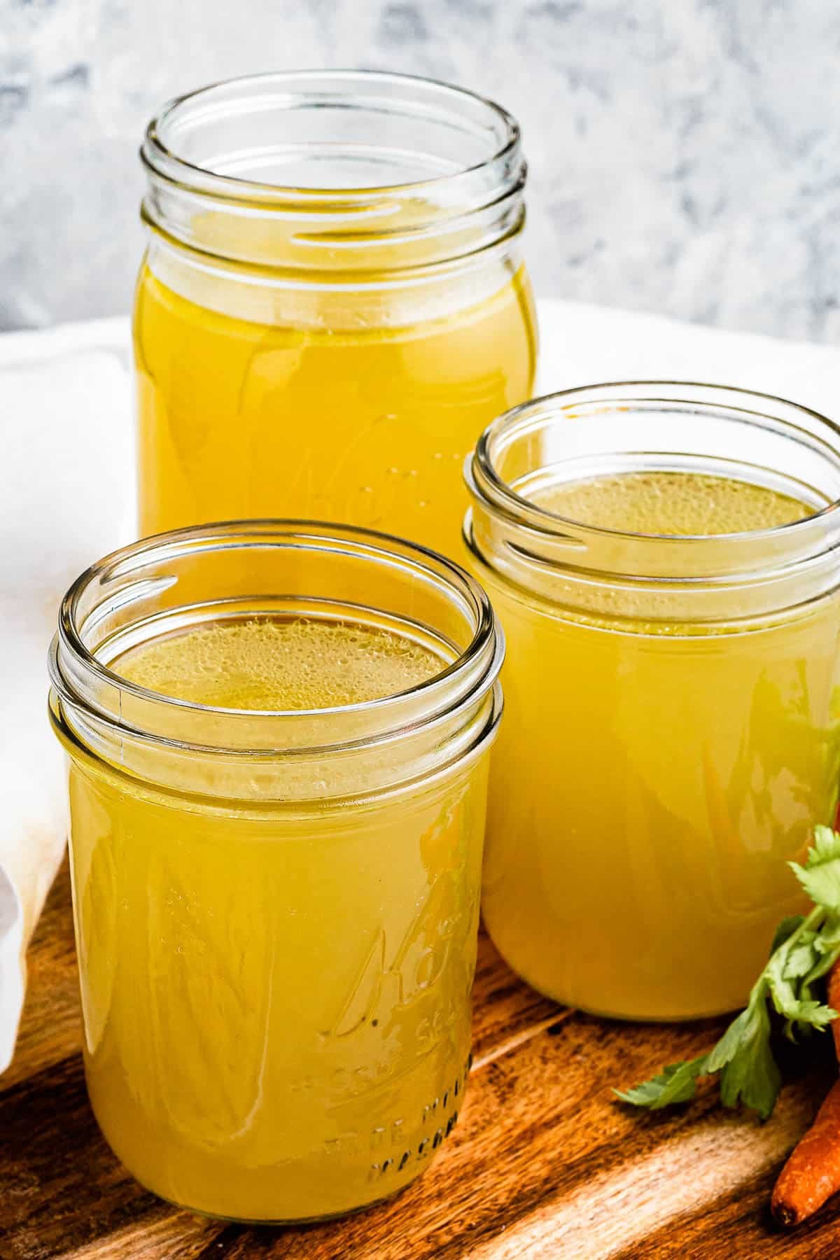 Three jars of chicken broth on a cutting board with raw carrots positioned next to them.