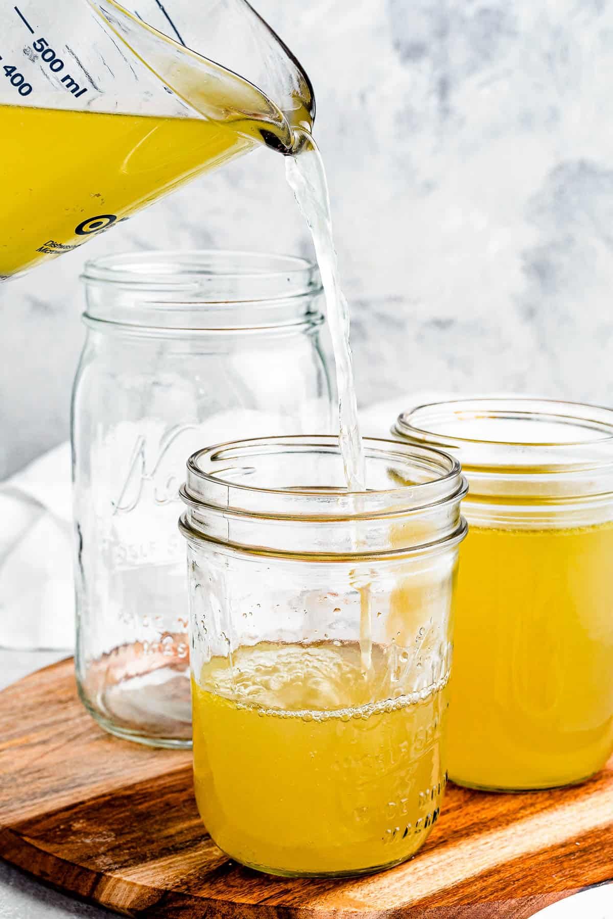 Chicken broth being poured from a large measuring cup into several jars.