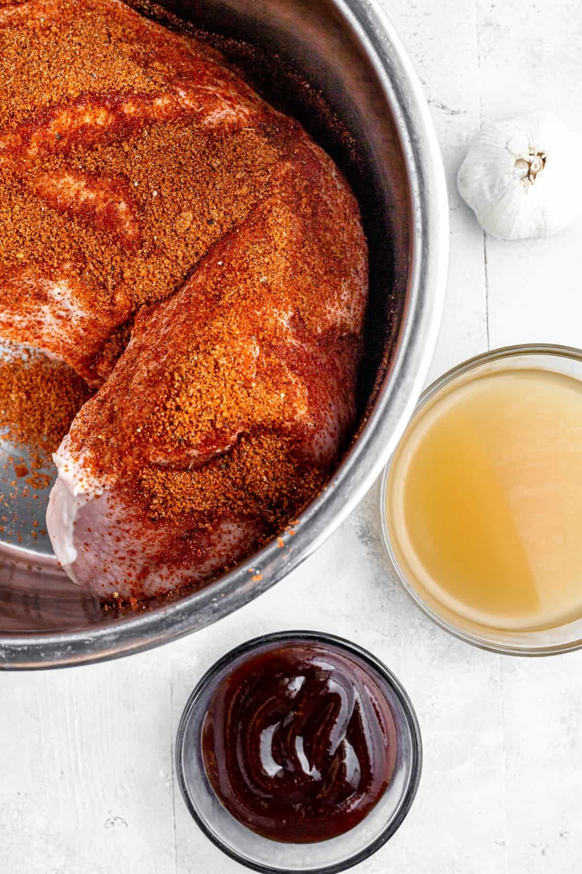 A season-rubbed pork loin in the metal insert of an Instant Pot, with a head of garlic, bowl of broth, and dish of barbecue sauce nearby on the work surface.