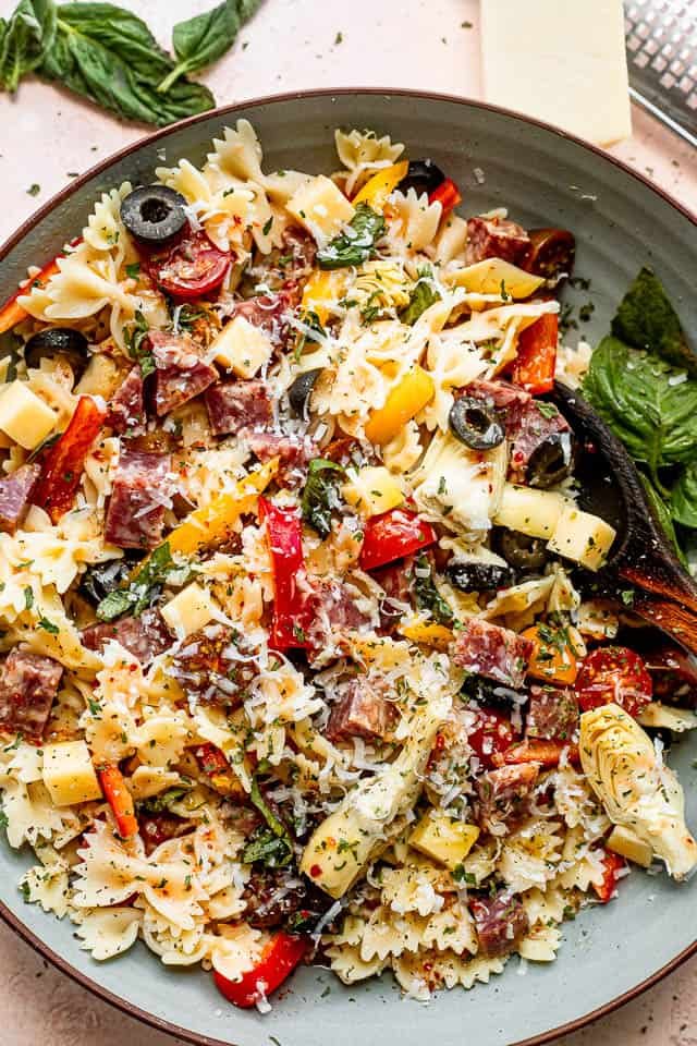 top view shot of a wooden spoon in a bowl filled with pasta, cheese cubes, salami cubes, tomatoes, and olives