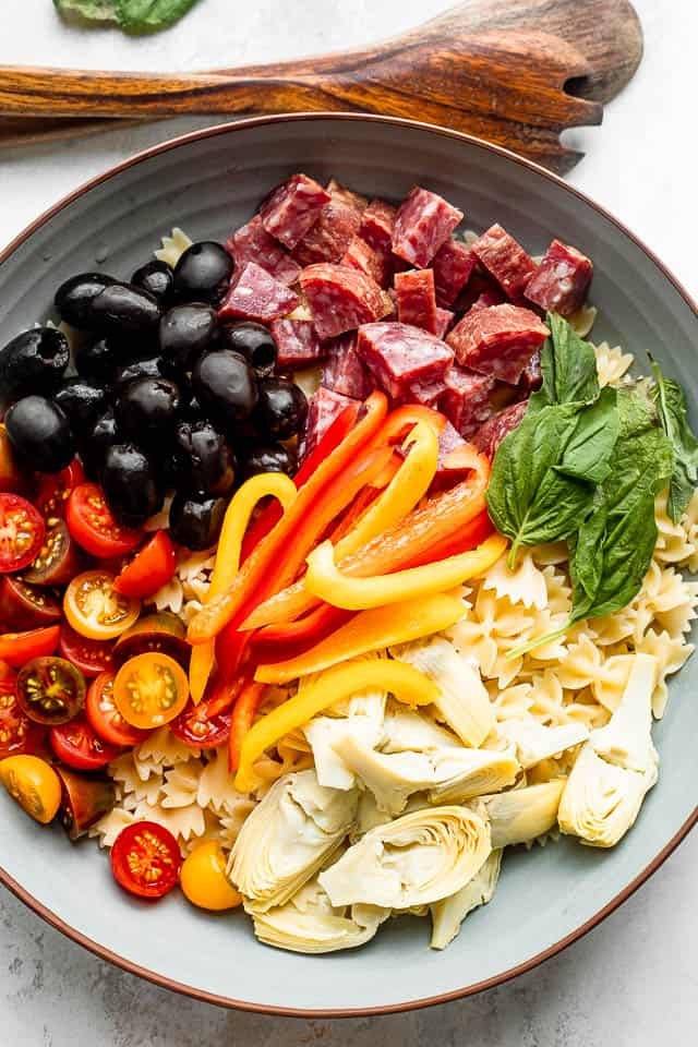 wooden spoons next to a salad bowl with arranged olives, salami, pasta, cheese, artichokes, tomatoes, basil leaves, and peppers