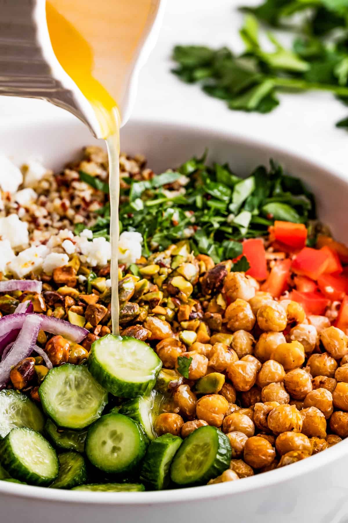 Pouring salad dressing over chickpeas, sliced cucumbers, red onions, peppers, herbs, and feta.