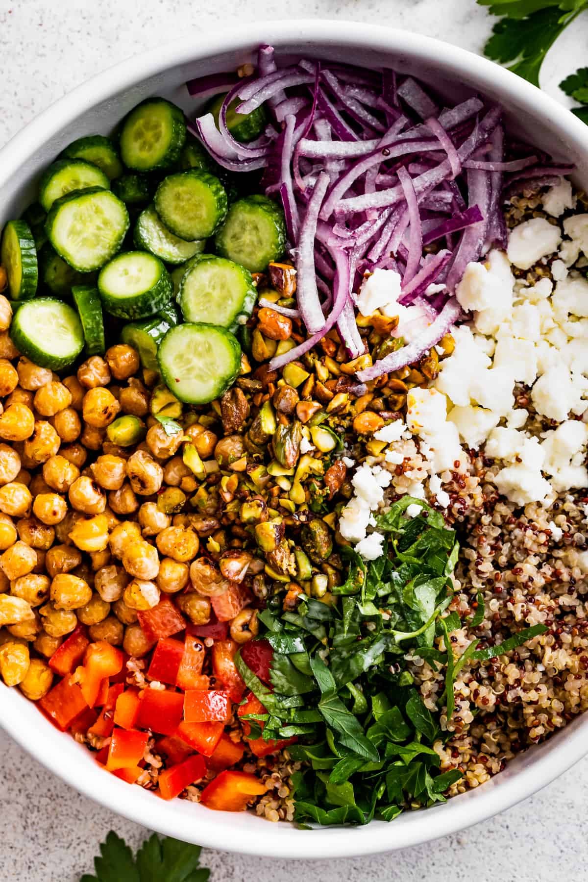 A large bowl with sliced cucumbers, sliced red onions, chickpeas, diced red bell peppers, herbs, tri-colored quinoa, and crumbles of feta cheese.