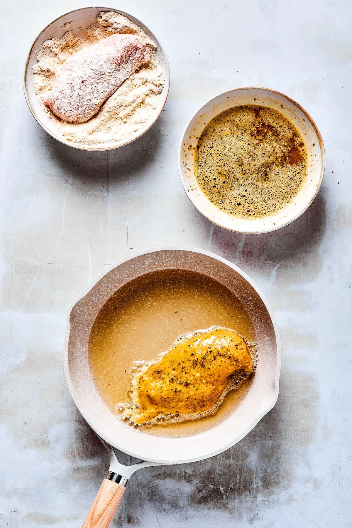 A chicken breast in a dish of dry breading ingredients, next to a dish of wet breading ingredients, and a skillet with one breaded chicken breast cooking in oil.