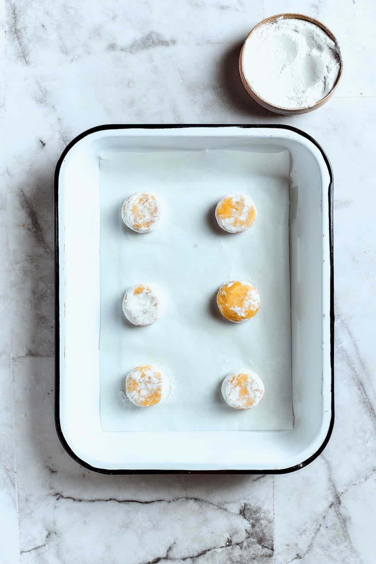 Overhead view of 6 balls of lemon cookie dough covered in powdered sugar in a baking tray.