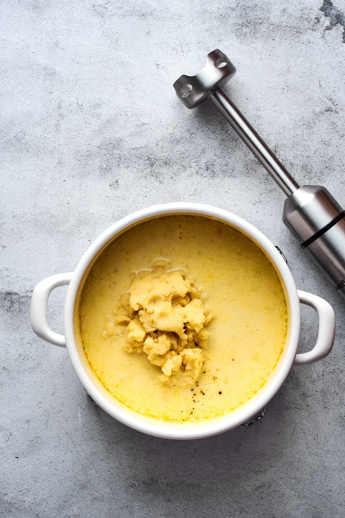 A soup pot with mashed potatoes and creamy soup inside. An immersion blender is nearby on the work surface.