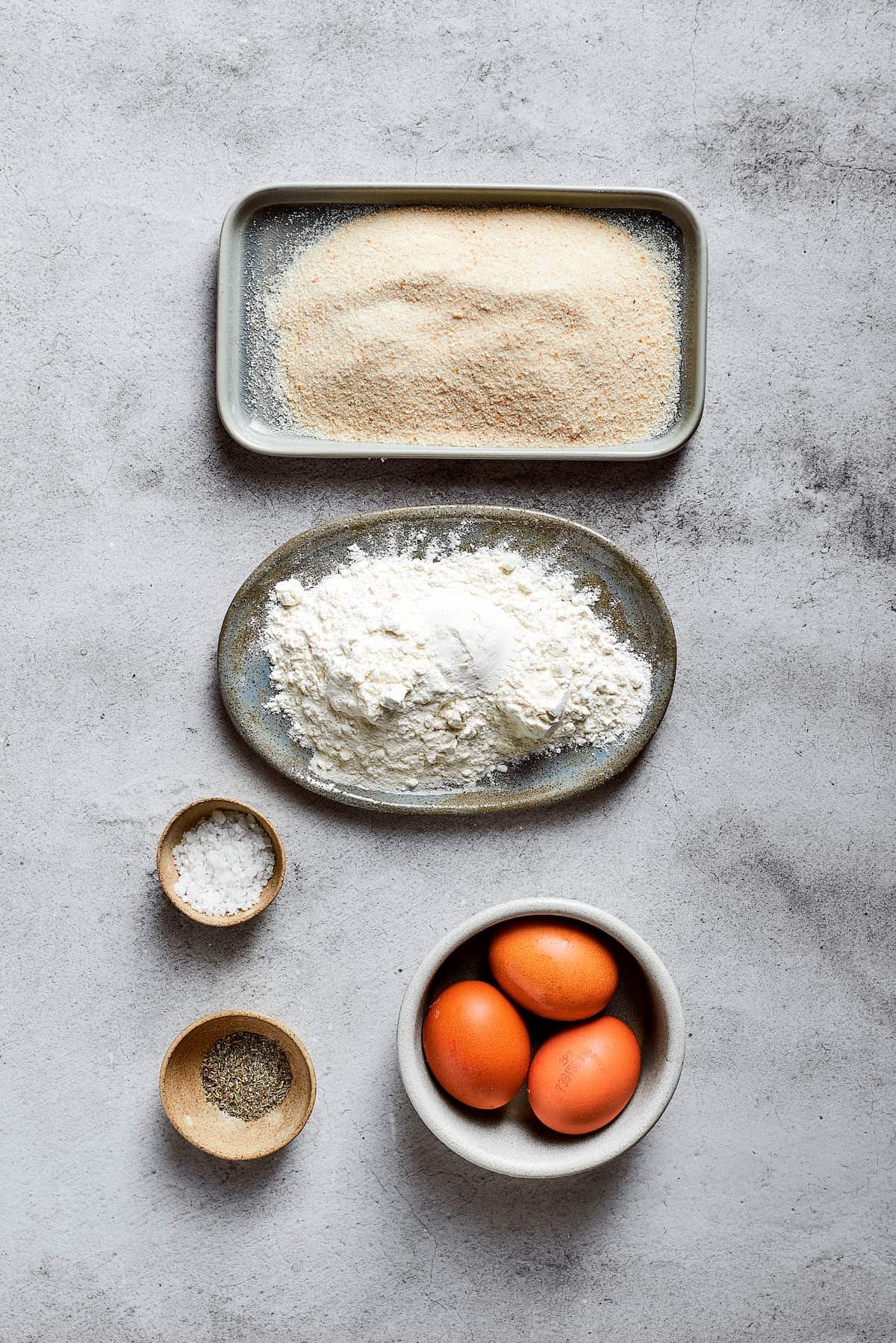 Overhead view of the ingredients needed for breading mac and cheese bites: a plate of flour, a plate of breadcrumbs, a bowl of salt, a bowl of Italian seasoning, and 3 eggs.