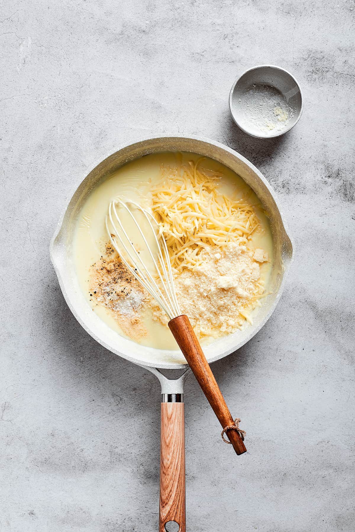 A sauce pan with a whisk and shredded cheese on top of milk.