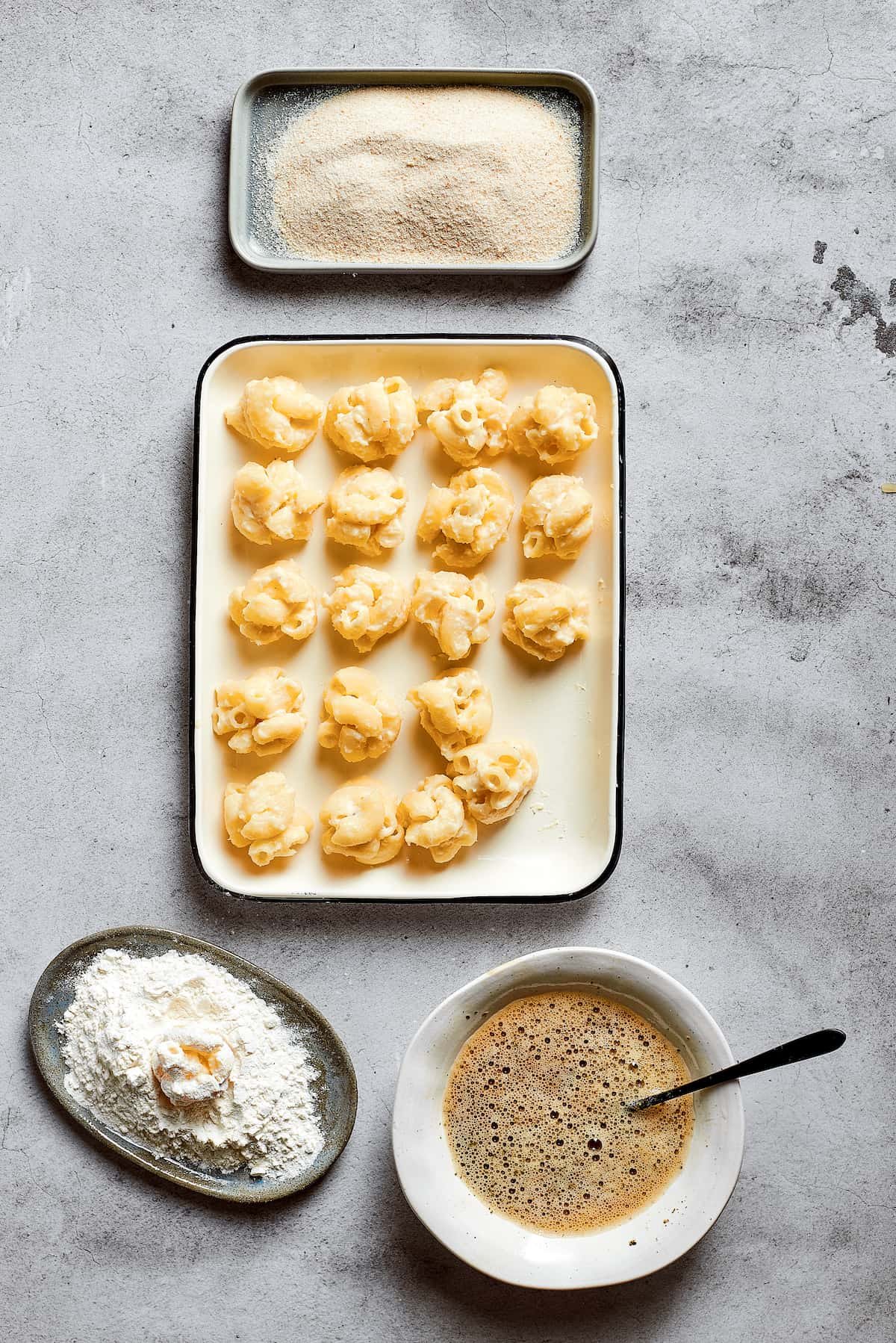 Overhead view of a tray of rolled balls of mac and cheese, next to a bowl of flour, a bowl of breadcrumbs, and a bowl of eggs beaten with Italian seasoning.