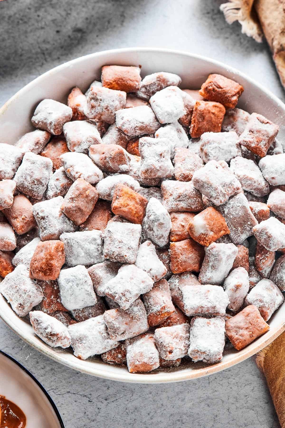 Close-up shot of puppy chow in a white bowl, to show the texture. Some pieces are fully coated in powdered sugar, while others are not.