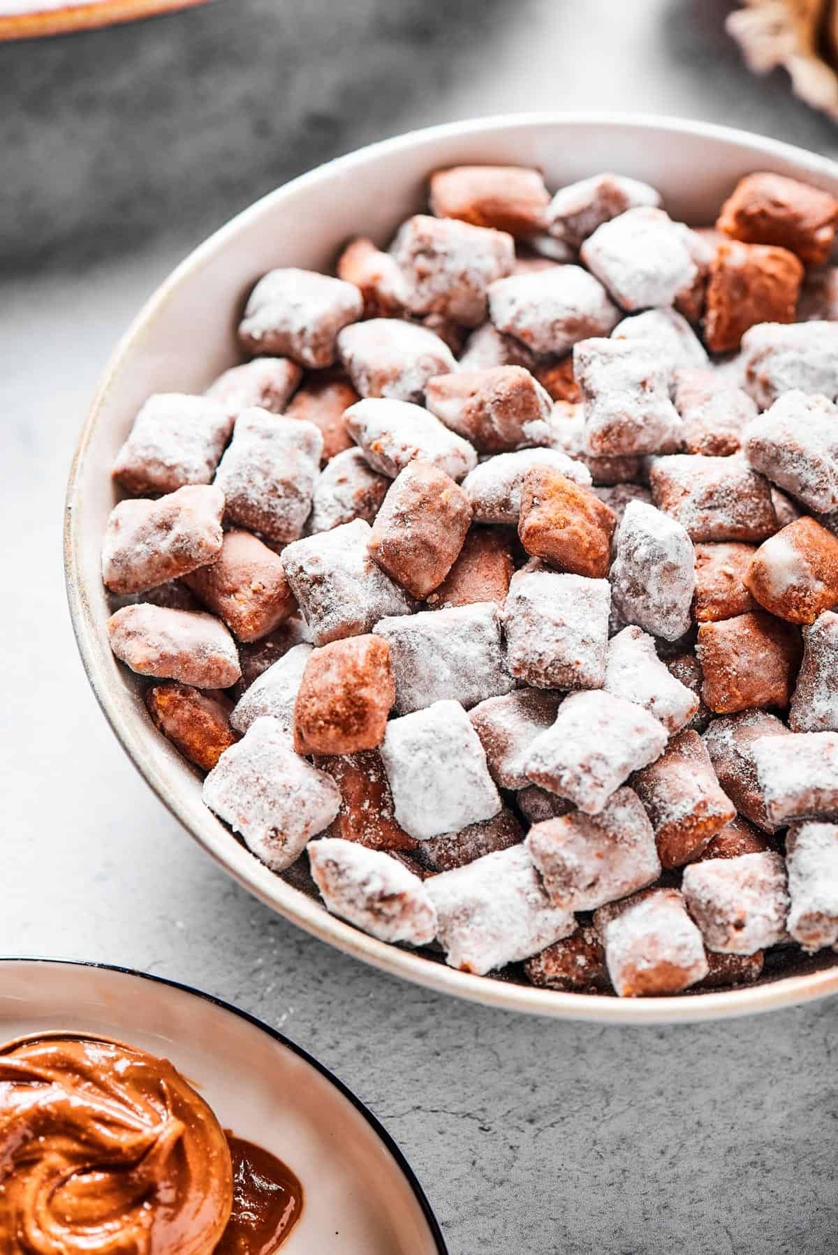 Close-up shot of puppy chow in a white bowl, to show the texture. Some pieces are fully coated in powdered sugar, while others are not.
