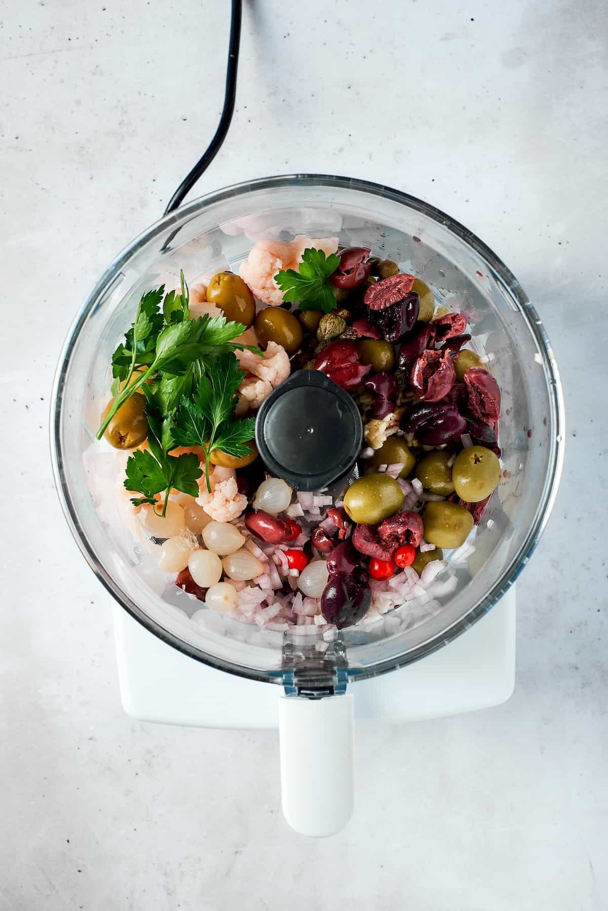 Olive salad ingredients are added to a food processor.