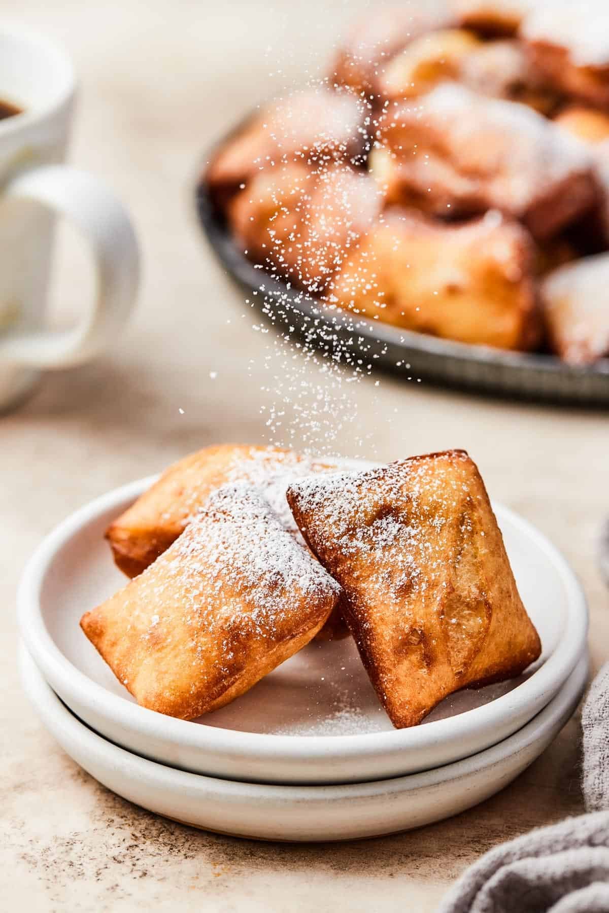 sprinkling powdered sugar over three beignets set on white plates.