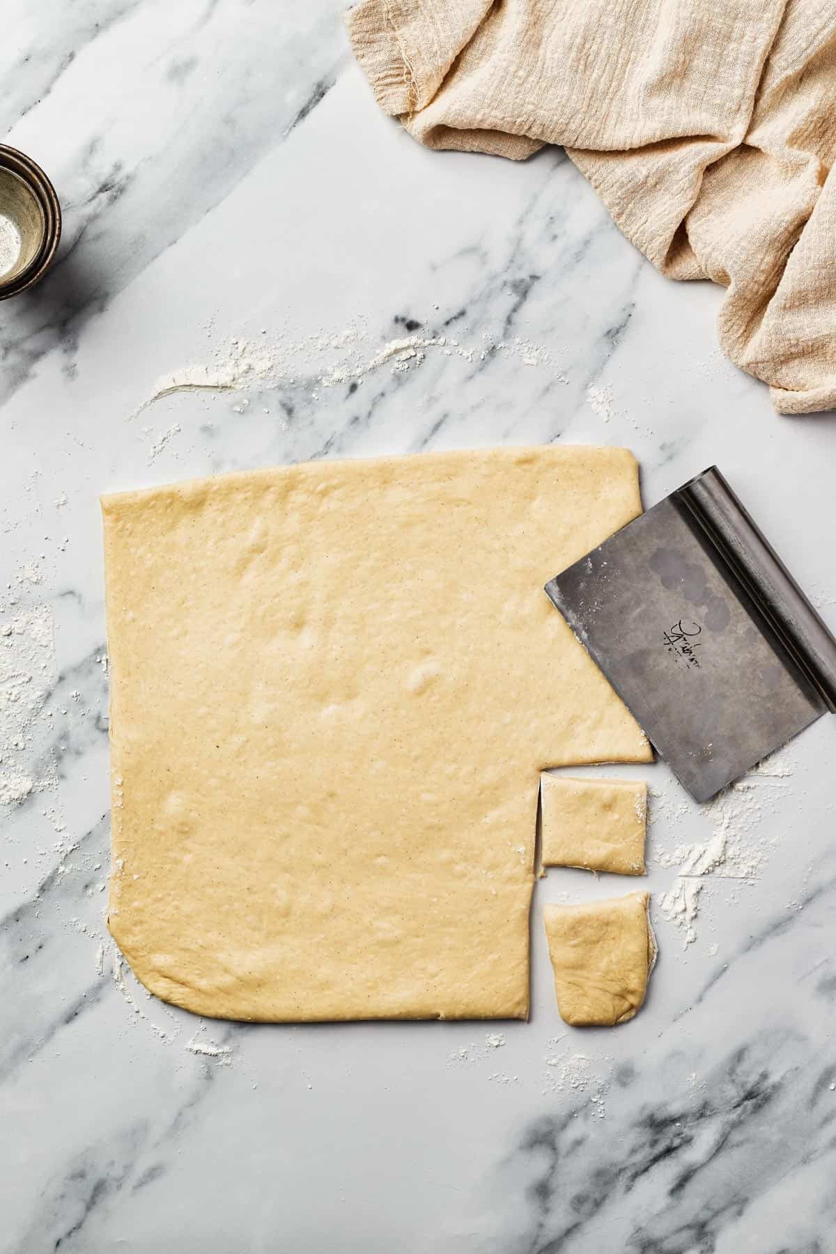 A square of rolled-out yeast dough on a marble work surface, with small square pieces cut out of it. A bench scraper is resting on the dough.