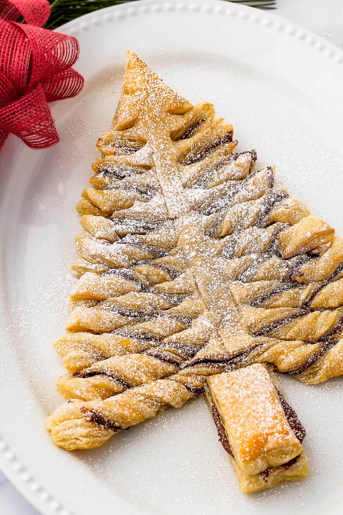 A baked puff pastry Christmas tree, dusted with powdered sugar, on an oval platter. Christmas greenery, a red bow, and a small bowl of chocolate dipping sauce surround the platter.