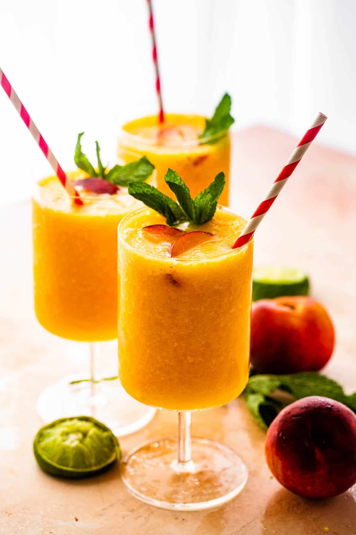 three glasses filled with peach daiquiris and garnished with mint, peach slices, and straws