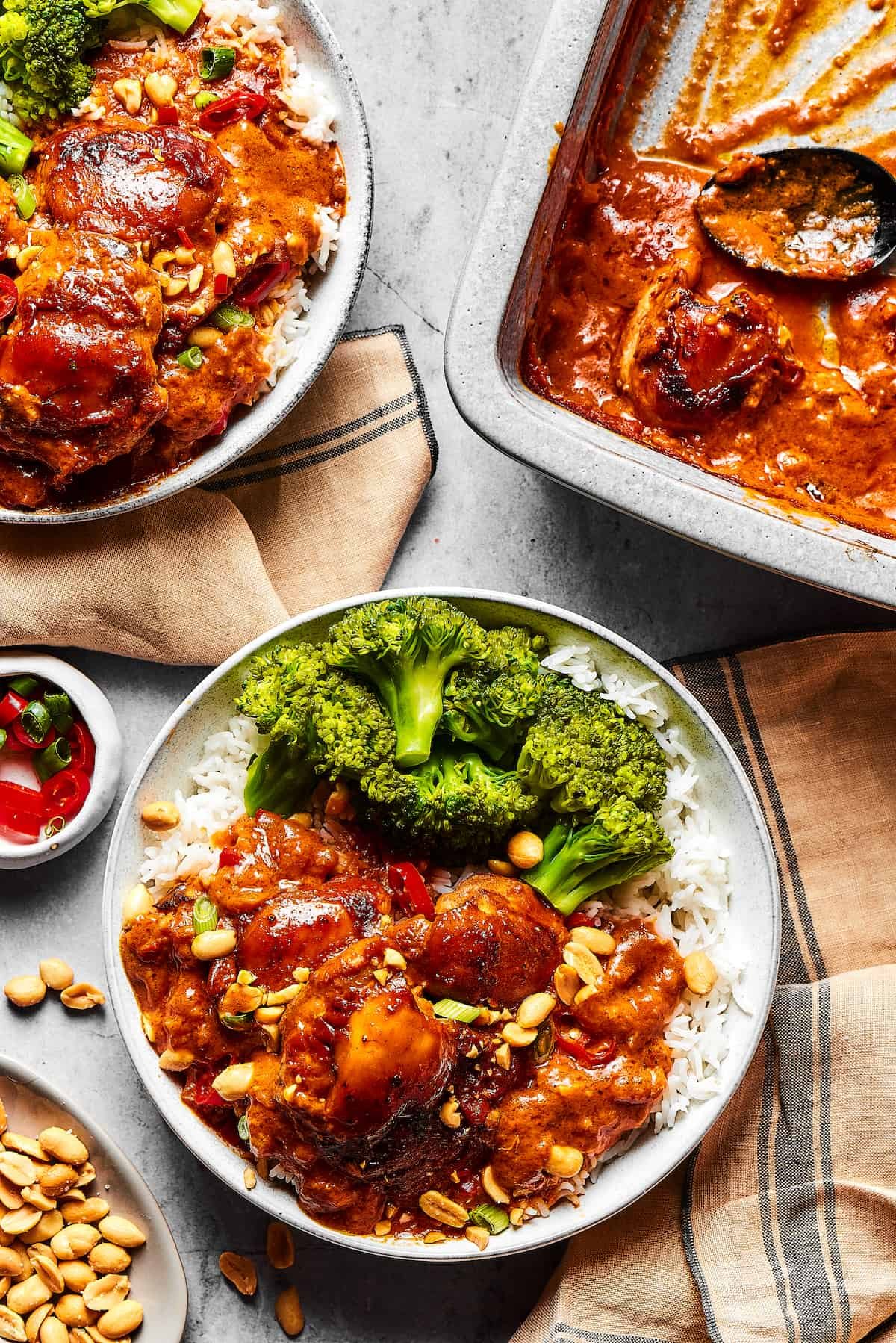 Peanut chicken served with broccoli over rice. A baking dish of the chicken is nearby on the table, along with garnishes and more plates.
