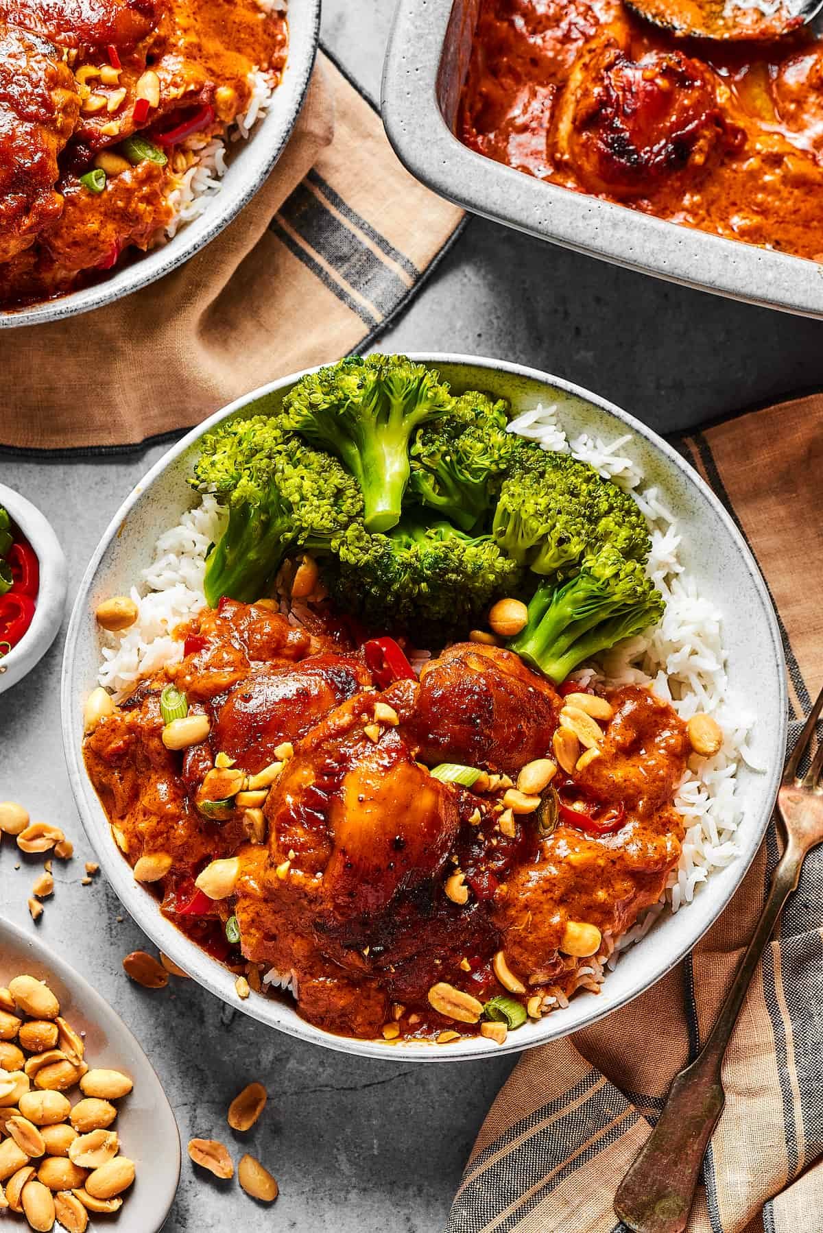 Peanut chicken served with broccoli over rice. A baking dish of the chicken is nearby on the table, along with garnishes and more plates.