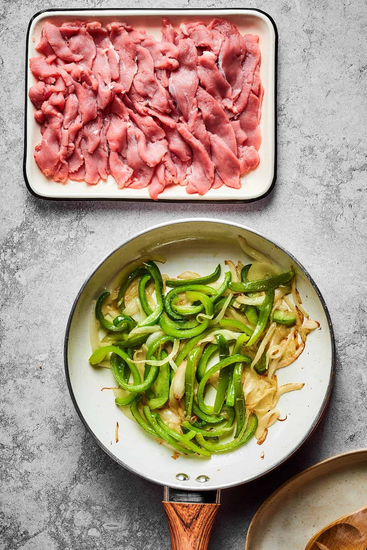 Overhead shot of a skillet of sauteing veggies, next to an enamel dish of sliced steak.