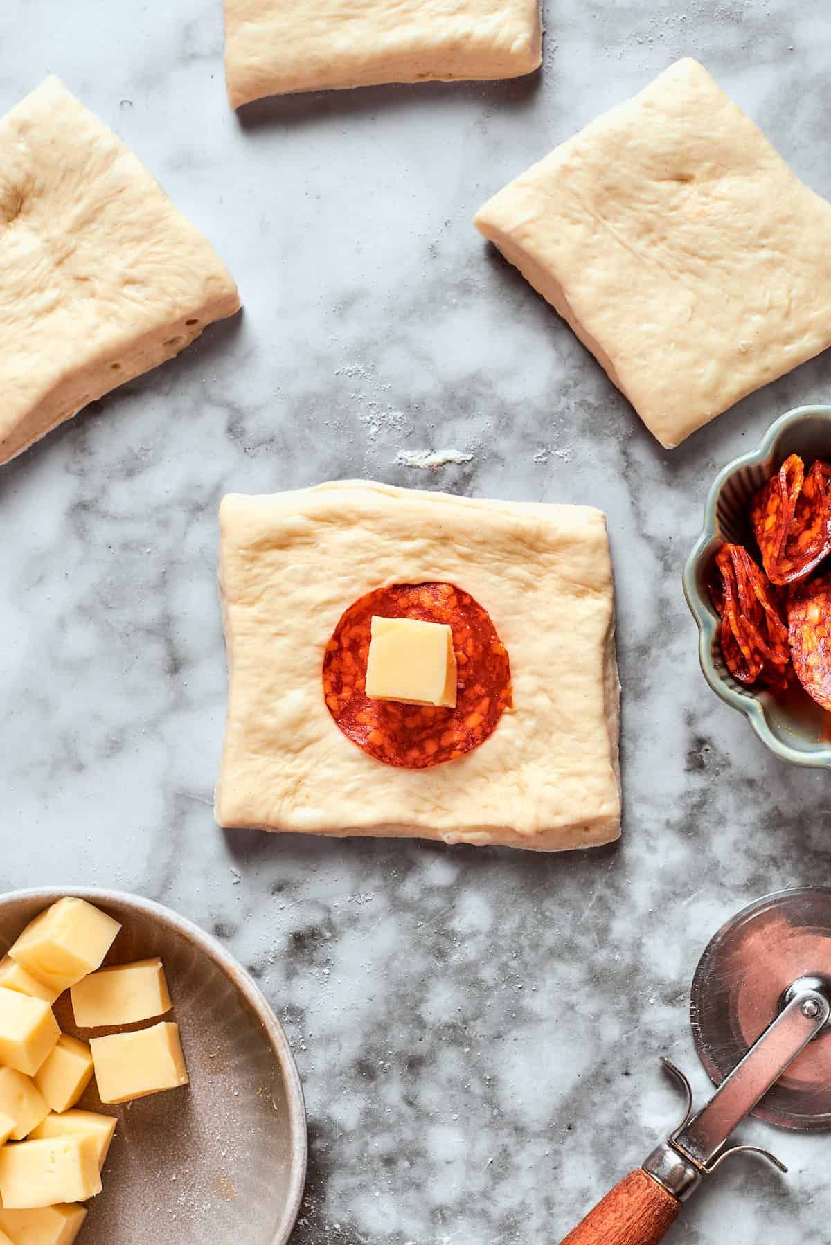 A slice of pepperoni and a cube of cheese is added to a square of dough.