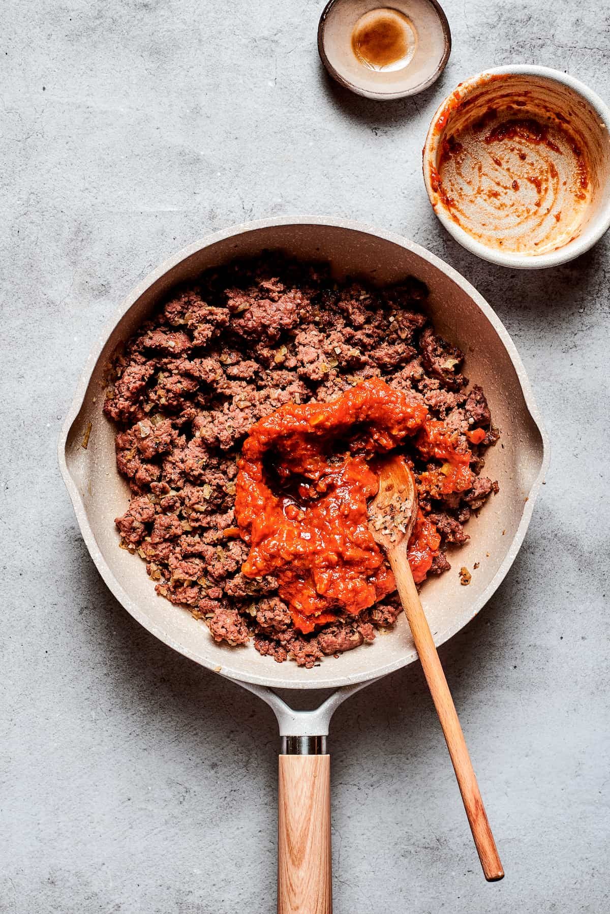 Pizza sauce is stirred into a pan of browned meat.