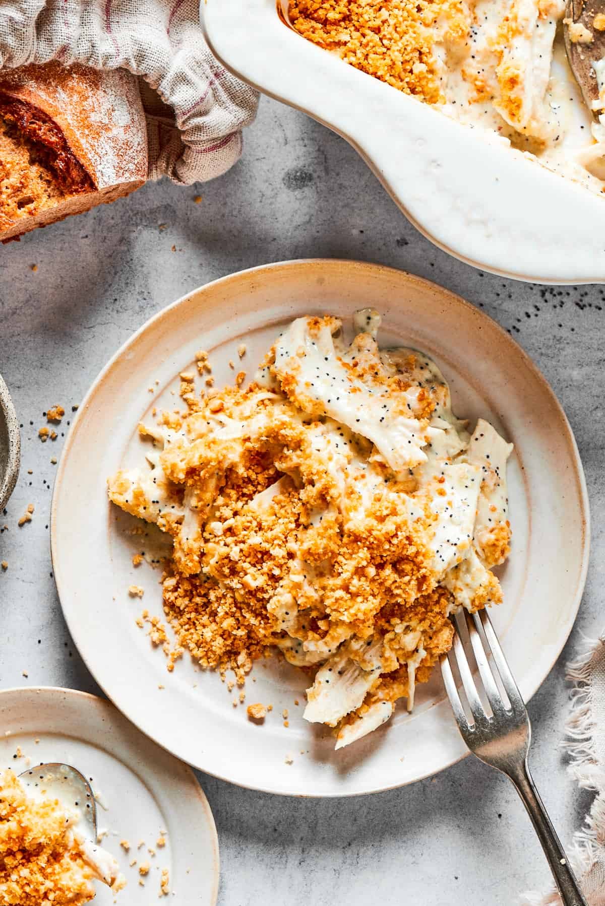 A casserole dish of poppy seed chicken is shown on a table with a loaf of bread and a plate holding a serving of the chicken.