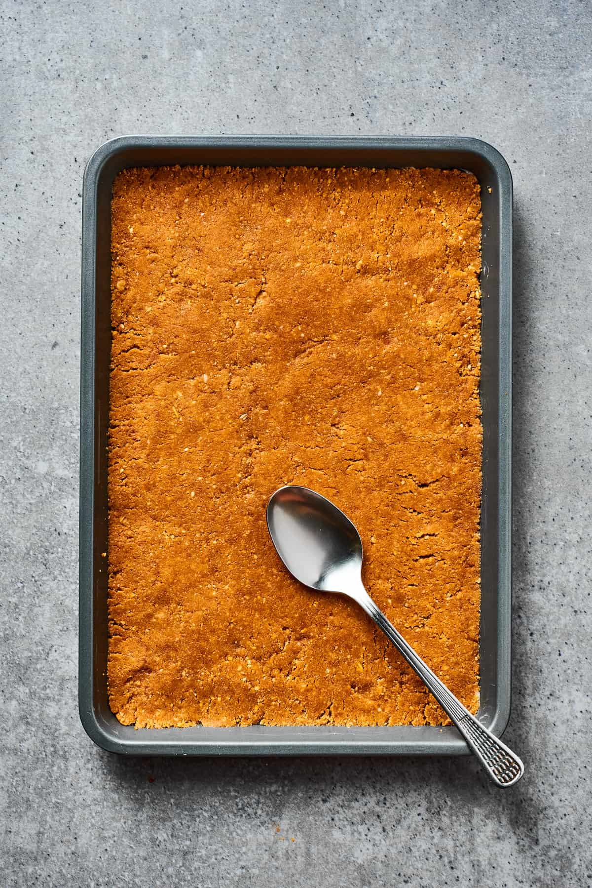 A cookie crust pressed into a baking dish. A spoon is resting on the crust.