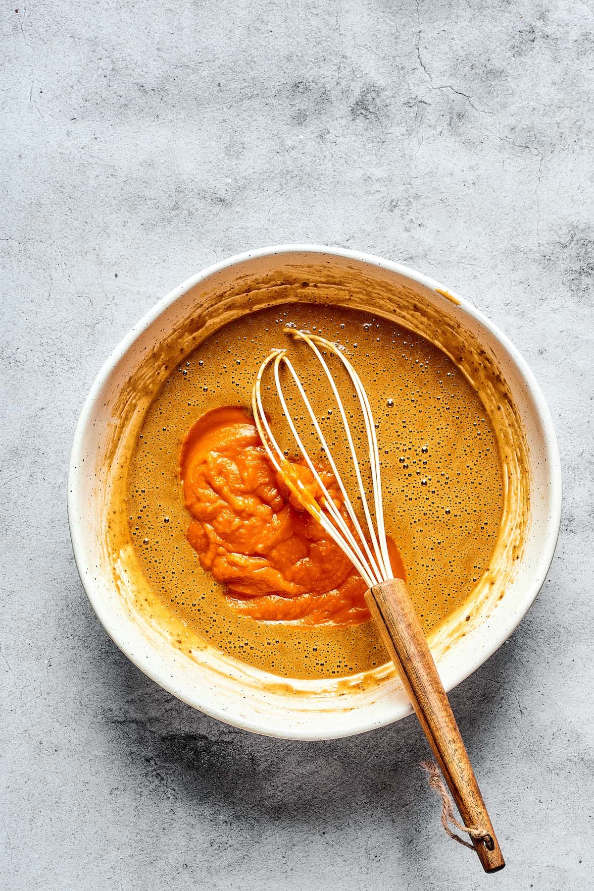 Wet muffin batter ingredients in a mixing bowl with a whisk, including pumpkin puree.