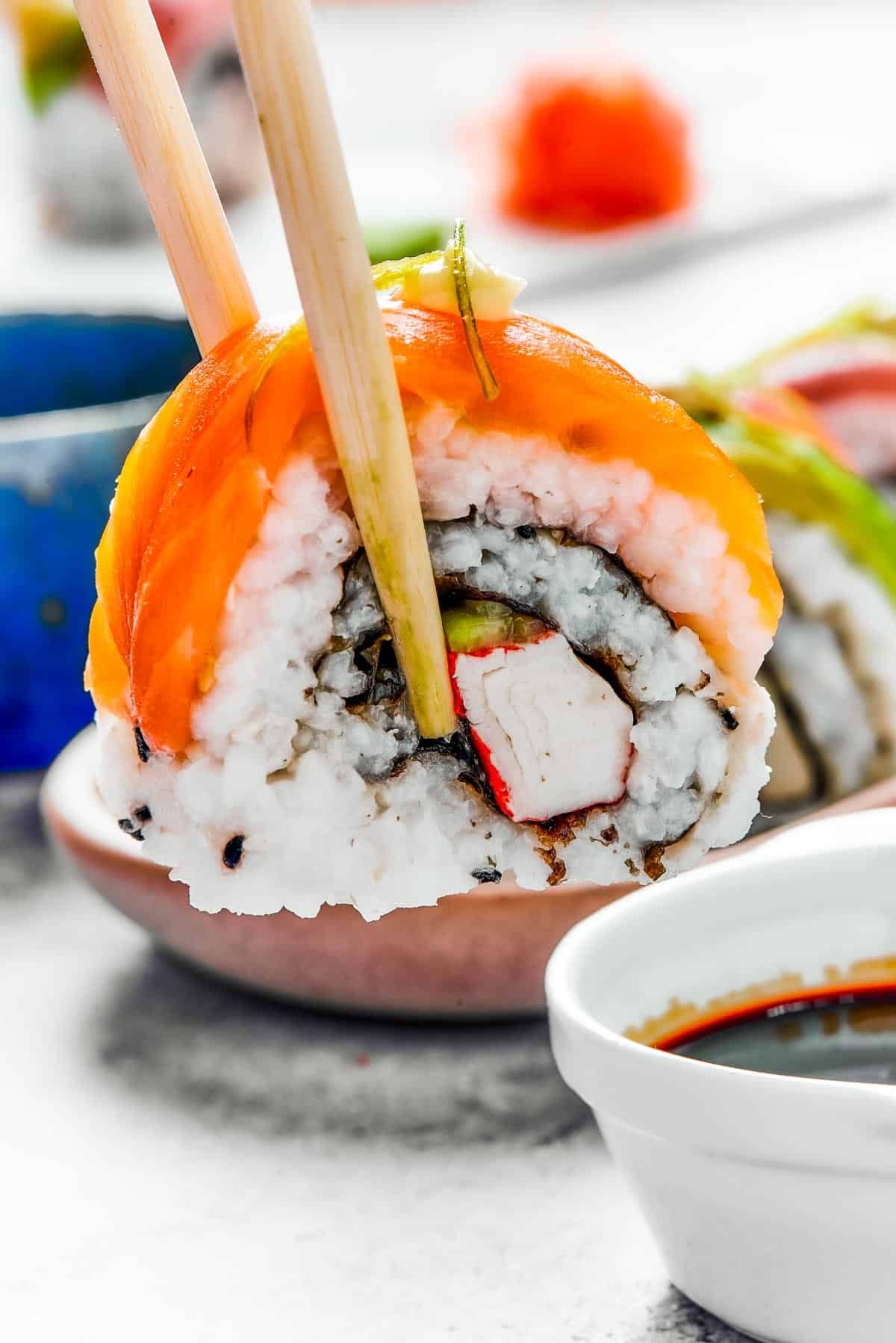 A bite of sushi held in a pair of chopsticks. A small plate of sushi and a container of soy sauce is in the background.