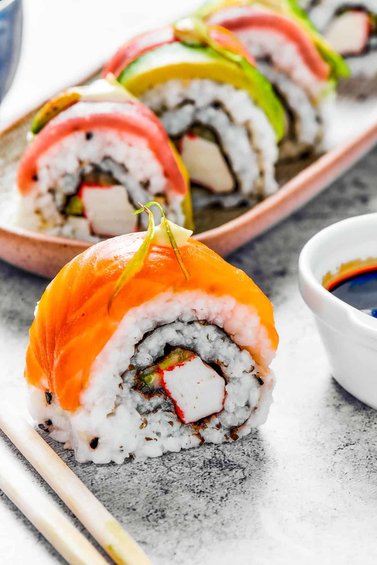 A bite of sushi set on a surface. A small plate of sushi and a container of soy sauce is in the background.