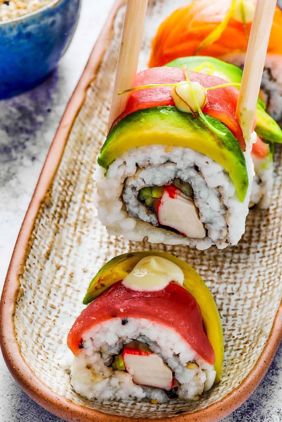 A bite of sushi held in a pair of chopsticks. A small plate of sushi and a container of soy sauce is in the background.