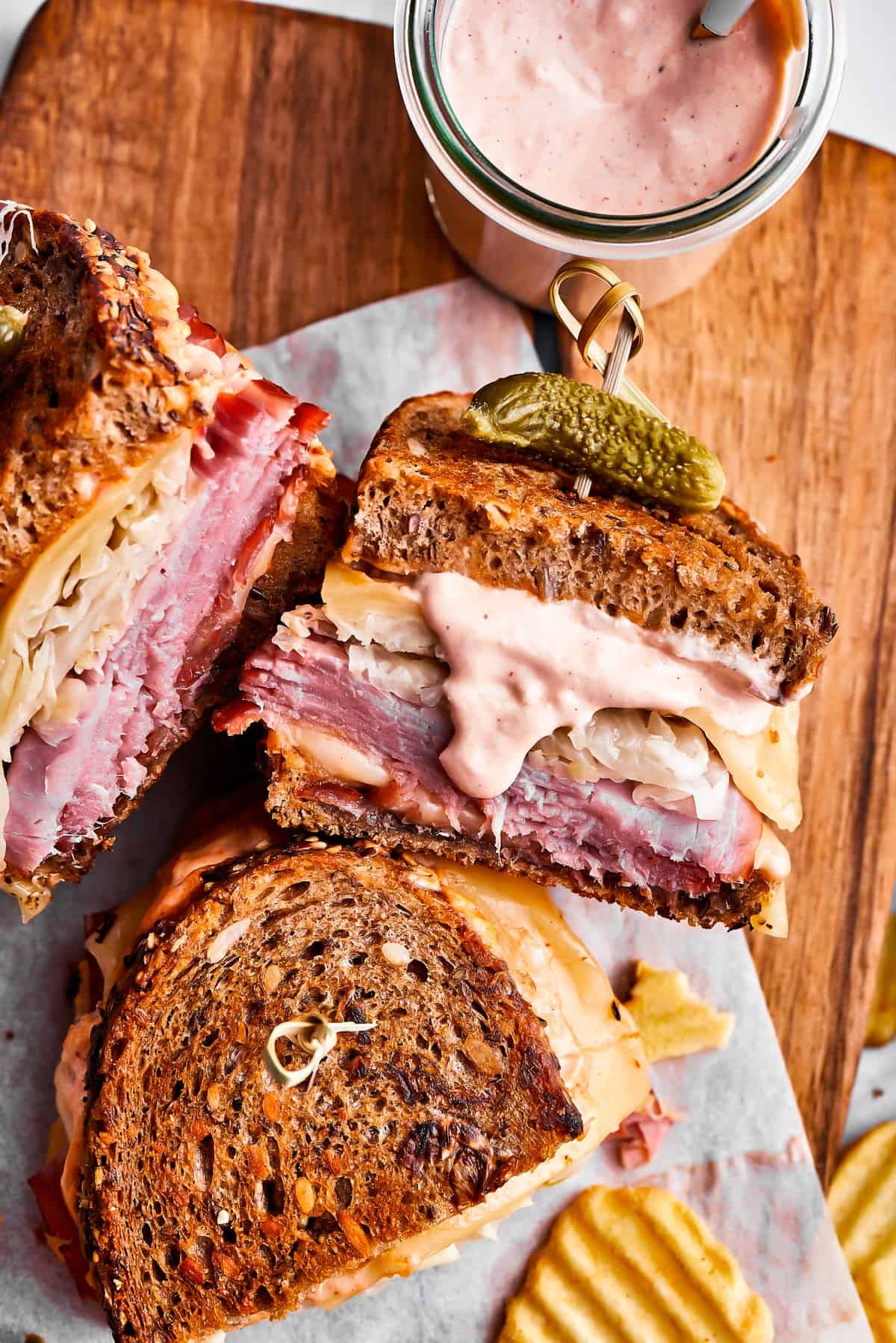 Three Reuben sandwich halves, some potato chips, and a jar of dressing on a cutting board.