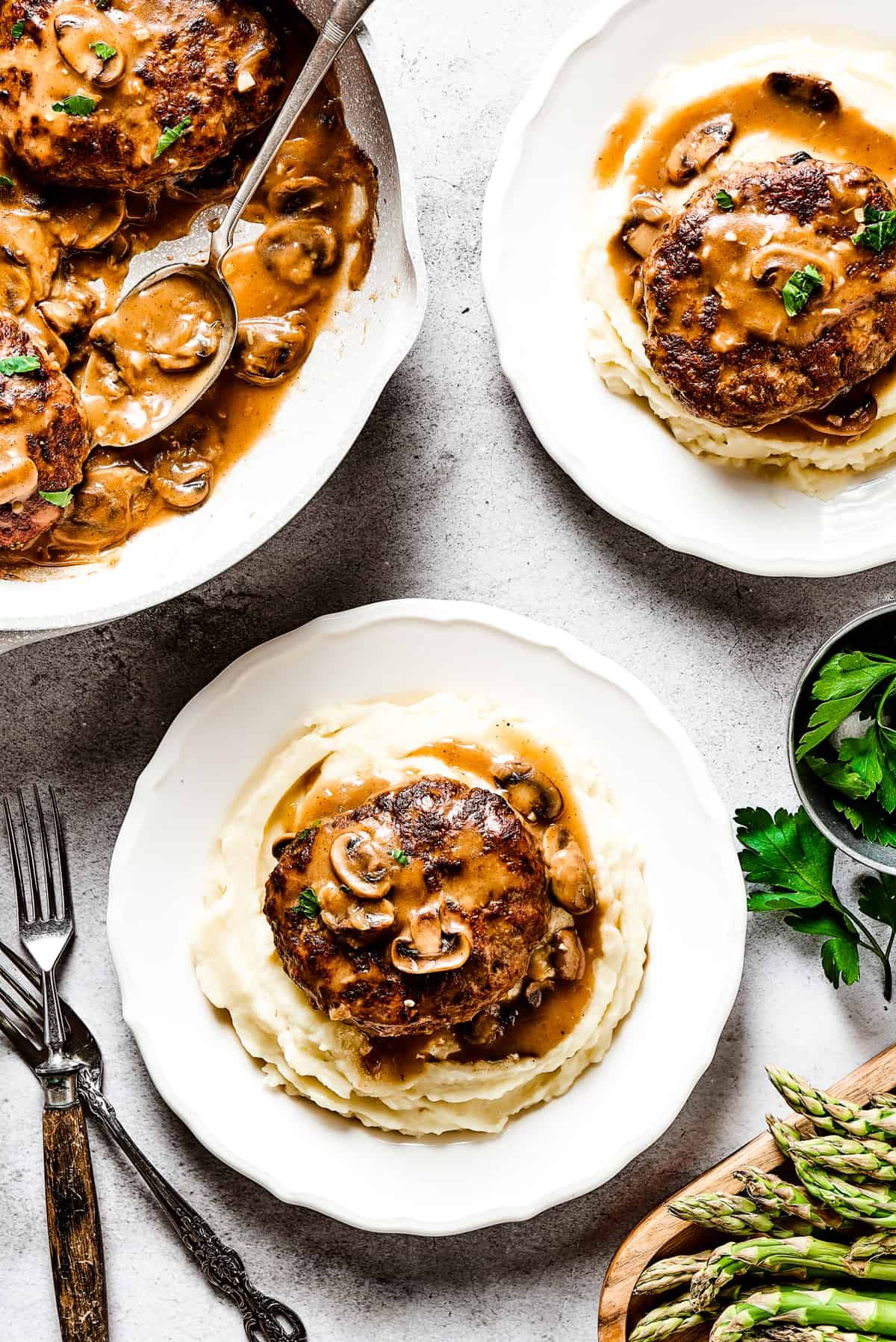 A skillet of beef patties in gravy next to two servings on dinner plates.